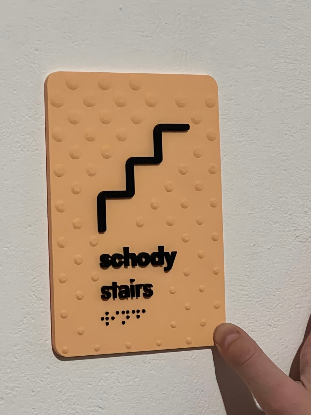 Dotykowa tabliczka kierunkowa schody z piktogramem i napisem w alfabecie Braille'a, trzymana w dłoni.